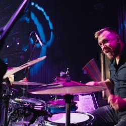 Person playing drums on a dark stage, lit up by stage lights