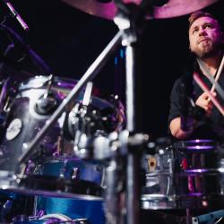 Person playing drums on a dark stage, lit up by stage lights